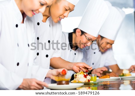 Asian Indonesian chef along with other cooks in restaurant or hotel kitchen cooking, finishing dish or plate for dessert