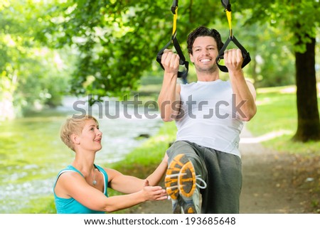 Young fitness man exercising with suspension trainer sling and personal sport trainer in City Park under summer trees