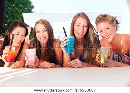 Four woman lying in the sand on the beach, drinking fancy cocktails and tanning in the sun