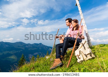 Hiking - Young couple sitting on mountain summit at cross in the Bavarian Alps and enjoys the panorama in the leisure time or in vacation