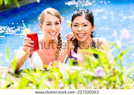 Two girls or women in vacation, Asian and Caucasian, in tropical garden bathing in hotel pool with drinks or cocktails