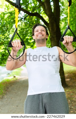 Young man exercising with suspension trainer sling in City Park under summer trees for sport fitness