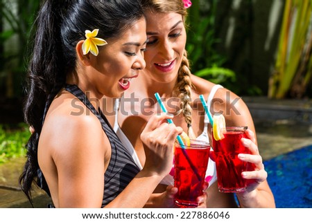 Two girls or women in vacation, Asian and Caucasian, in tropical garden at hotel pool tanning being served drinks or cocktails