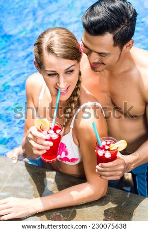 Couple in vacation, Asian man and Caucasian woman, in tropical garden bathing in hotel pool with drinks or cocktails
