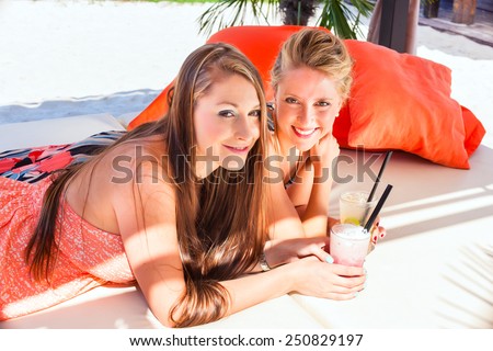 Two woman lying in the sand on the beach, drinking fancy cocktails and tanning in the sun