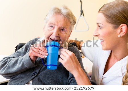 Nurse in nursing home giving drink to elderly woman sitting in wheelchair