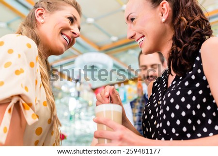Two woman friends in coffee bar drinking latte macchiato in glass, a barista preparing drinks in the background