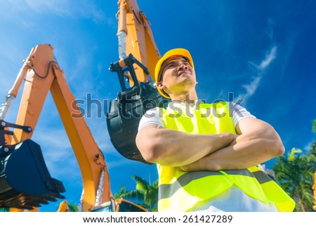 Asian worker standing in front of construction machinery of building site or rental company