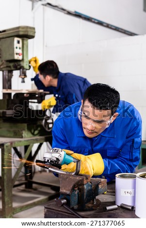 Two Asian industrial workers in metal factory with electrical grinding tool and power drill machine