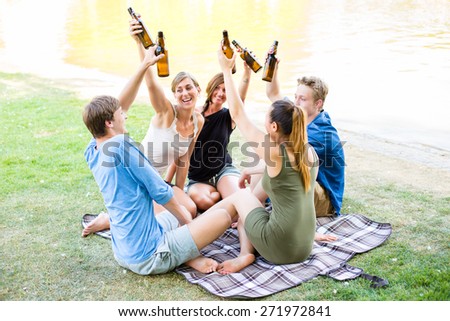 Students in park toasting with beer bottles having picnic at river