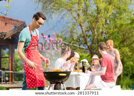 Family and friends having bbq at garden party, man in the foreground on grill, in background people drinking and eating