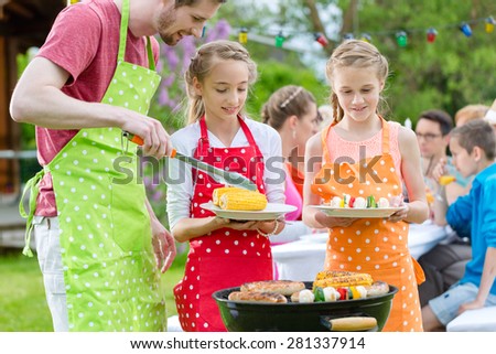 Family having barbecue at garden party, dad is putting corn and grilled vegetables on plates of daughters