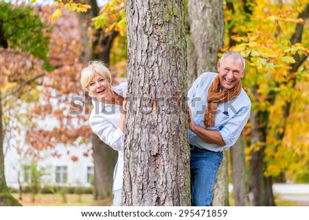 Couple, senior man and woman, flirting with each other playing hide and seek around a tree in fall tree