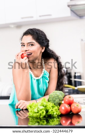 Indian woman eating healthy apple in her kitchen, salad and vegetables on the counter