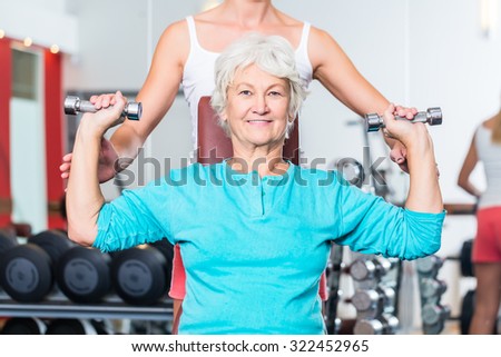 Senior women with fitness trainer in gym lifting dumbbell for pectoral training as sport exercise