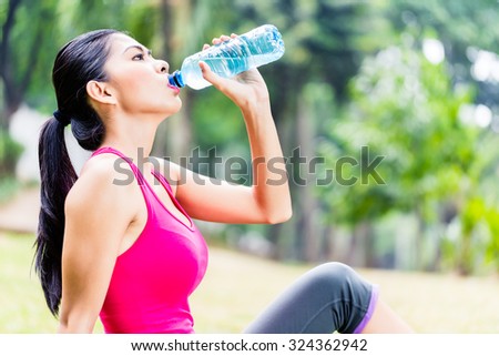 Asian woman having break from sport training in tropical park, drinking water from a bottle