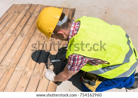 High-angle view of a blue-collar worker wearing yellow hard hat, safety vest and gloves while using a hammer during work on the construction site