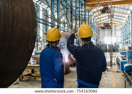 Two Asian experts talking while supervising the fabrication of industrial boilers indoors