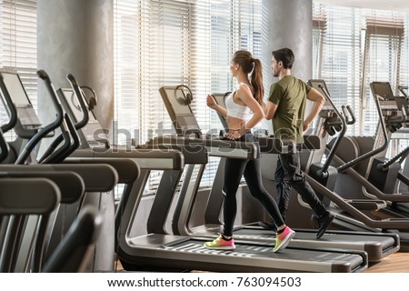 Side view of fit young man and woman smiling while running side by side on modern electric treadmills at the gym