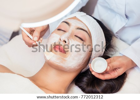 Close-up of the face of a young woman, relaxing under the gentle touch of the specialist applying on her cheeks white facial mask with rejuvenating effects