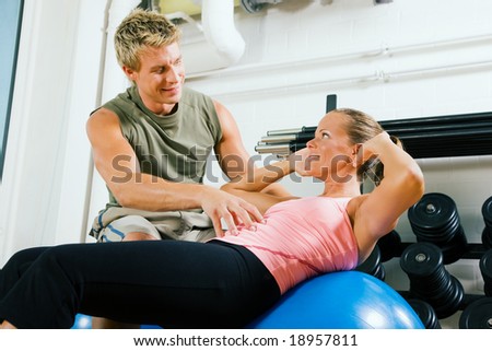 Woman doing sit-ups on a blue fitness ball in the gym, assisted by her personal trainer
