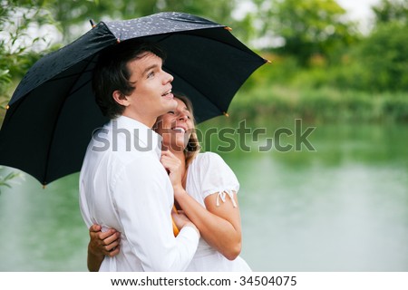Couple (man and woman) at a lake in the rain with an umbrella, laughing and having fun despite of the bad weather