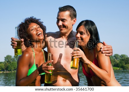 Group of friends - one man hugs two women and all have drinks in swimwear on the beach of a lake in summer