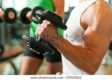 Strong man exercising with dumbbells in a gym, in the background a woman also lifting weights; focus on hands