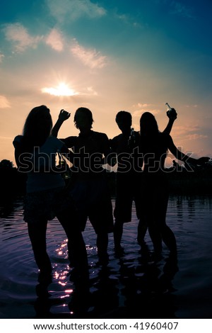 People (two couples) on the beach dancing to music, drinking and having a lot of fun in the sunset (only silhouette of people to be seen, people having bottles in their hands)