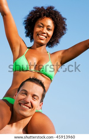 Couple in love - Woman of Brazilian origin in bikini sitting on her man\'s shoulders under blue sky - summer and fun