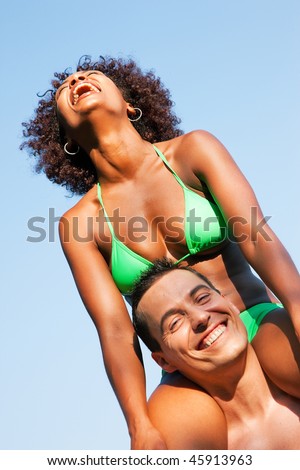 Couple in love - Woman of Brazilian origin in bikini sitting on her man\'s shoulders under blue sky - summer and fun