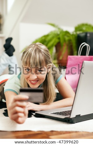 Woman lying in her home living room on floor shopping or doing banking transactions online in the Internet, emphasized by shopping bags in the background and her holding a credit card