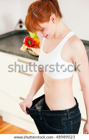Woman trying her last year\'s jeans finding them way too big after she lost so much weight; in the background a fruit bowl to be seen as a symbol for healthy eating