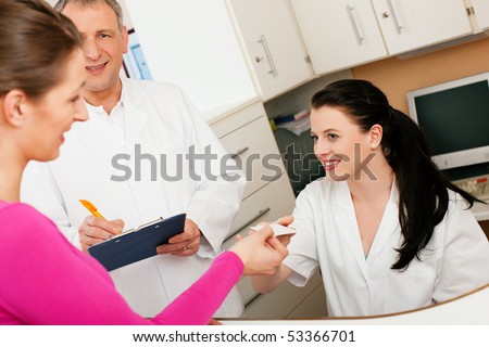 Patient in reception area of office of doctor or dentist, handing her health insurance card over the counter to the nurse, the doctor standing in the background and is writing things on a clipboard
