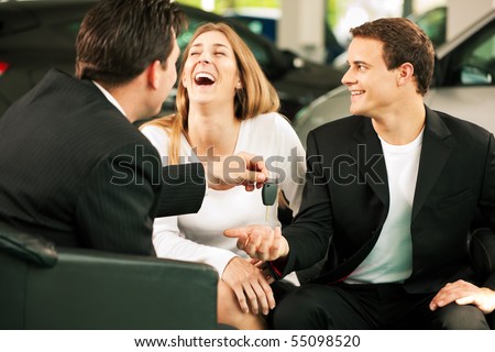 Sales situation in a car dealership, the dealer is handing auto keys to a young couple, they are excited, cars standing in the background