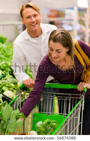 Man and woman in a supermarket at the vegetable shelf shopping for groceries, she is putting some stuff into the shopping cart