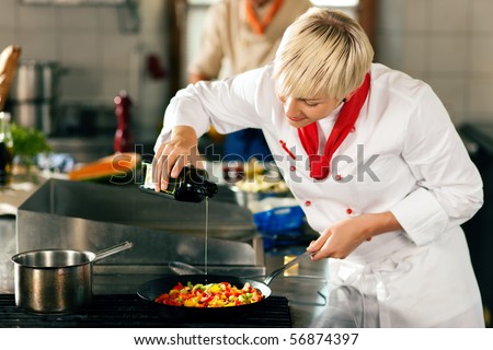 Two chefs in teamwork - man and woman - in a restaurant or hotel kitchen cooking delicious food, she is putting olive oil in the ratatouille