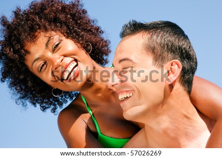 Couple in love - bikini-clad woman of color hugs a Caucasian man from behind under clear blue sky, both in beachwear in summer