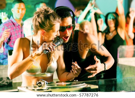 Two DJs - male and female, black and white - in a club at the turntable, in the background a crowd of their fans cheering