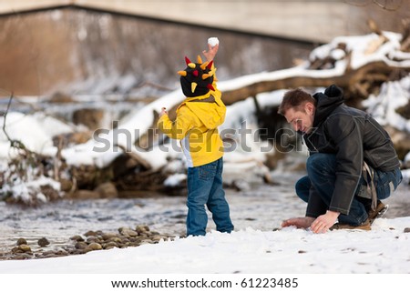Family - father and son to be seen - on a walk along a riverbank in winter; the child is throwing a snowball
