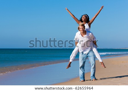 Couple in love - Caucasian man having his African-American woman piggyback on his back under a blue sky on a beach, she is stretching her arms