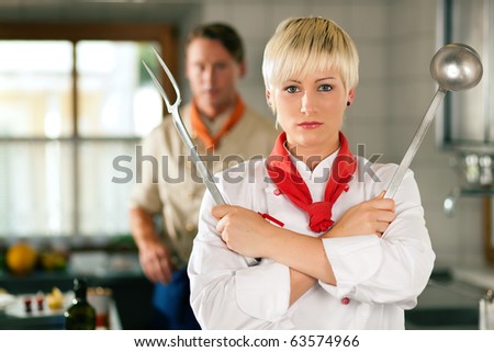 Female Chef in a restaurant or hotel kitchen posing with kitchen gear, in the background another chef