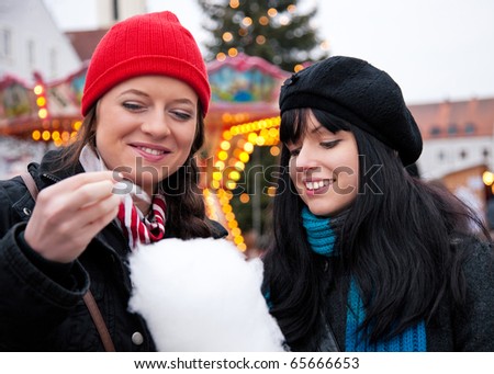 Two women on Christmas market eating cotton candy in front of a booth, it is cold
