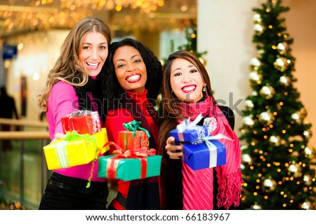 Group of three women - white, black and Asian - with Christmas presents in a shopping mall in front of a Christmas tree