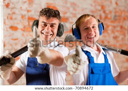 Two construction workers standing in a construction site in front of a brick wall. Their are wearing ear protection and having the thumps up