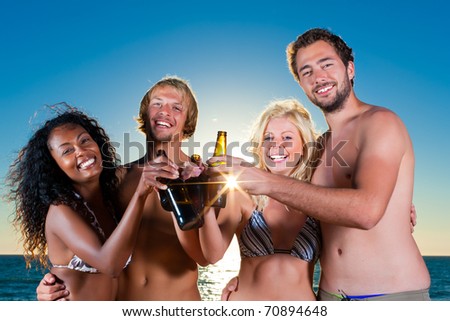 Group of four friends - men and women - standing with drinks on the beach against the setting sun over the ocean