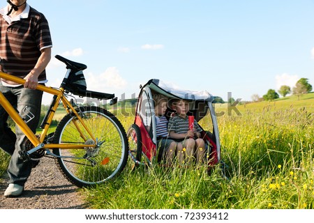 Dad driving his two children on a weekend excursion with bikes on a summer day in beautiful landscape, for safety and protection they are sitting in a bike trailer