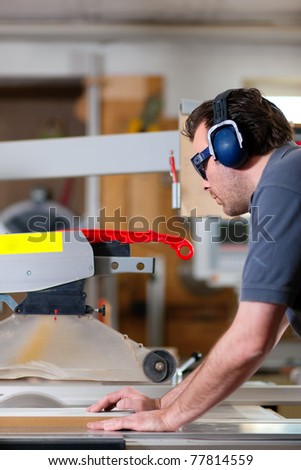 Carpenter working on an electric buzz saw cutting some boards, he is wearing safety glasses and hearing protection to make things safe
