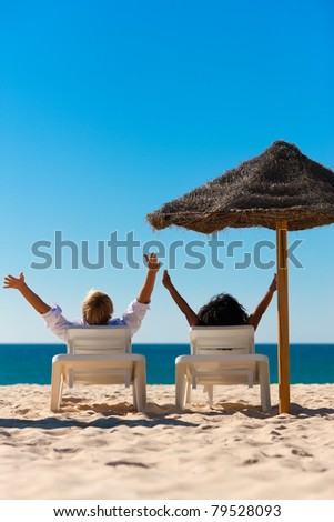 Couple sitting in sun chairs under an parasol sunshade on a beach stretching arms, feeling free