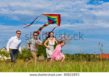 Happy family - mother, father, children - running over a green meadow in summer; they fly a kite